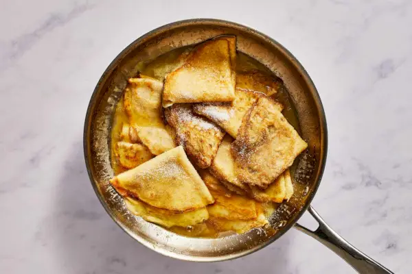 Eine große Pfanne mit gefalteten Crêpes, die in Butter-Orangen-Sauce gekocht und mit Zucker garniert werden