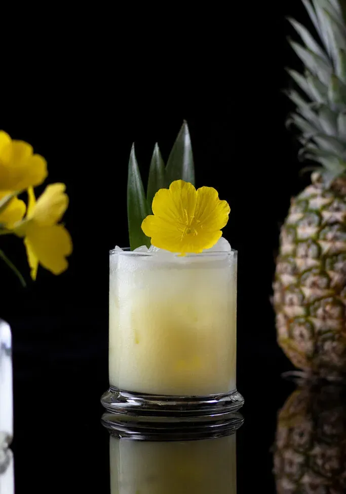 una bebida de color amarillo pálido en un vaso bajo adornado con una flor amarilla y hojas de piña.' title=