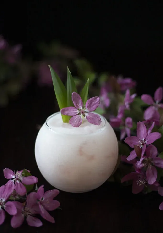 una bebida de color rosa pálido en un vaso redondo adornado con flores de manzano rosa.' title=