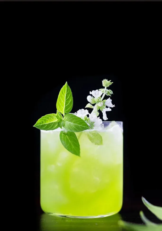 una bebida de color verde brillante en un vaso bajo con una flor de albahaca blanca.' title=