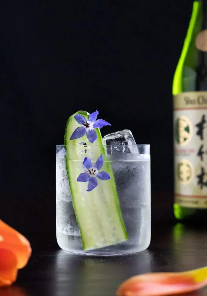una bebida clara en un vaso bajo con una rodaja de pepino y flores de borraja azul.' title=