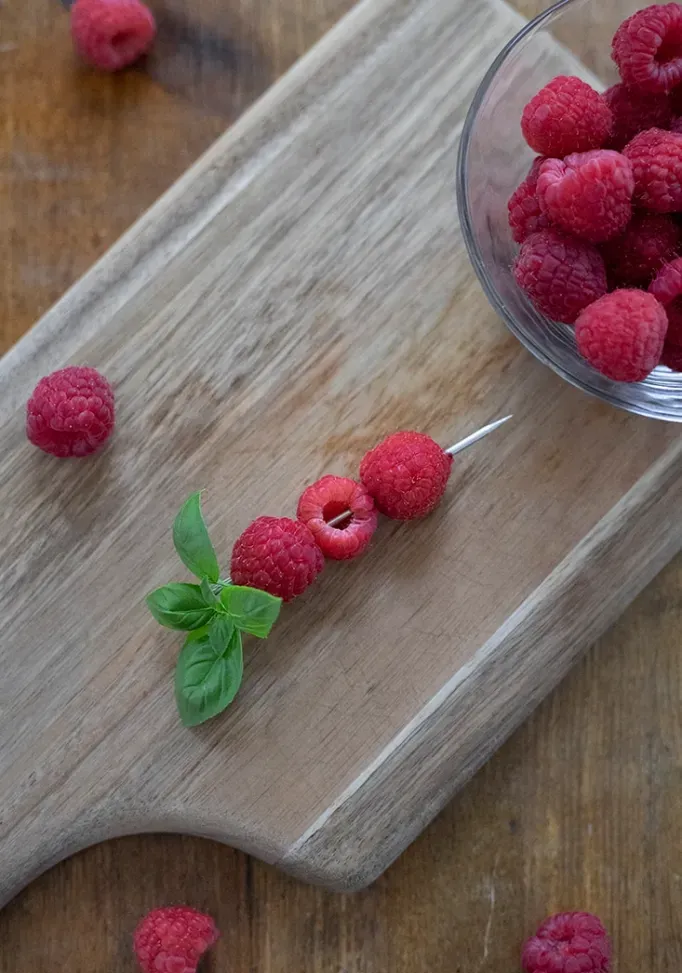 שלושה פטל משופדים על מקט קוקטייל עם ענף בזיליקום על קרש חיתוך מוקף בפטל' loading='lazy' title=