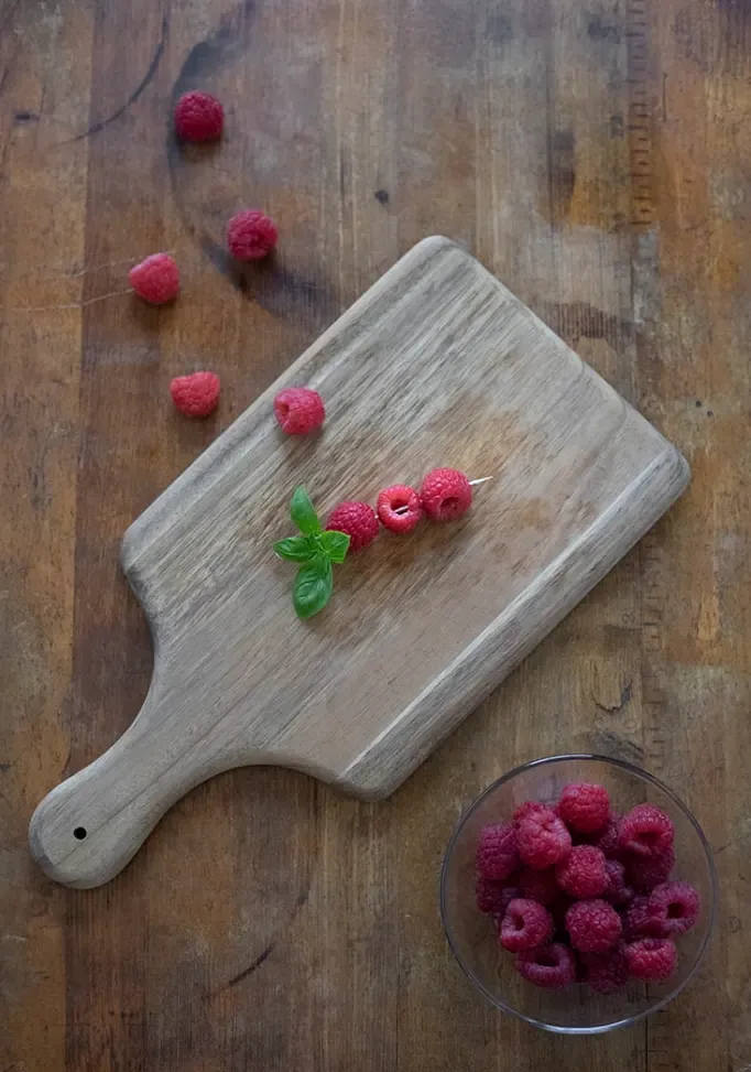 מבט מלמעלה למטה של ​​קרש חיתוך על שולחן עץ ישן עם מבחר קוקטייל אחד ועליו שלושה פטל משופדים וענף עלי בזיליקום' title=