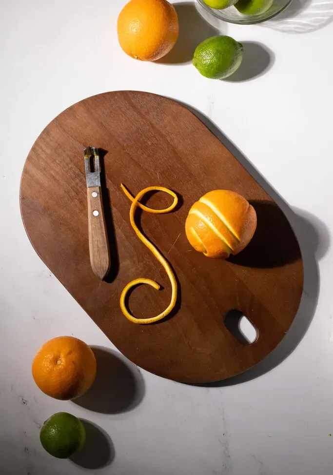 un trozo largo y fino de piel de naranja junto a una naranja y un cuchillo canal.' title=