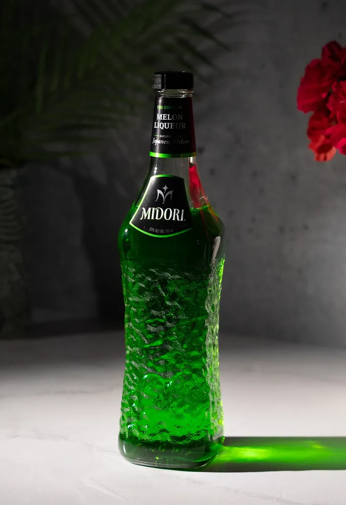bottiglia di liquore al melone Midori verde brillante accanto a fiori rosa.' loading='lazy' title=
