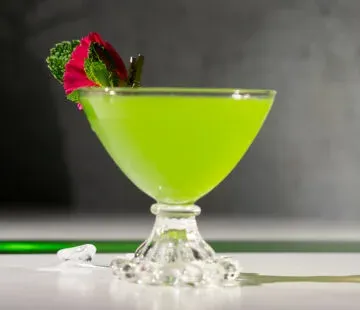 Cocktail verde neon in un bicchiere coupé corto con foglie di menta e un petalo di fiore rosa sul bordo.' title=