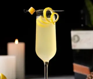 eine Champagnerflöte, gefüllt mit einem sprudelnden gelben Getränk, garniert mit einer Zitronenschale.' title=