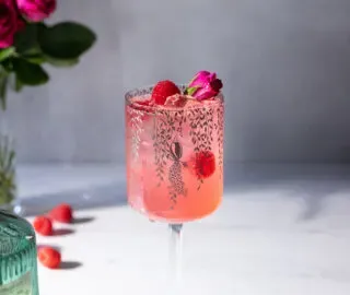 un bicchiere di vino alto pieno di una bevanda rosa con lamponi e una rosa.' title=