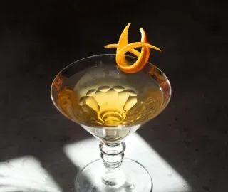 un cóctel de color dorado en una copa de martini con un toque de naranja.' title=