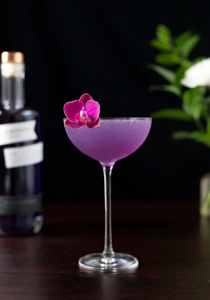 Lila Cocktail in einem Coupé-Glas neben der Empress Gin-Flasche.' loading='lazy' title=