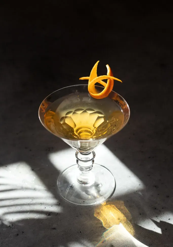 un cóctel de color dorado en una copa de martini con un toque de naranja.' title=