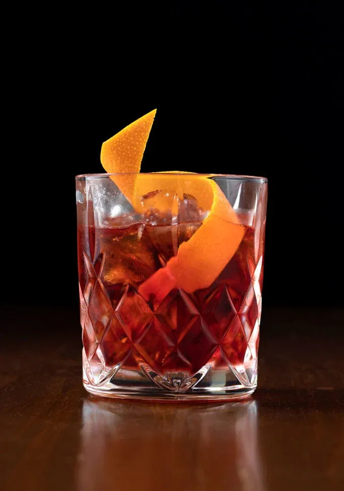 una bebida roja con un toque de naranja en un vaso bajo grabado sobre un fondo oscuro.' title=