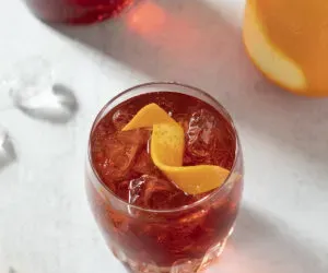 un cóctel de color rojo brillante en un vaso bajo con piel de naranja.' title=