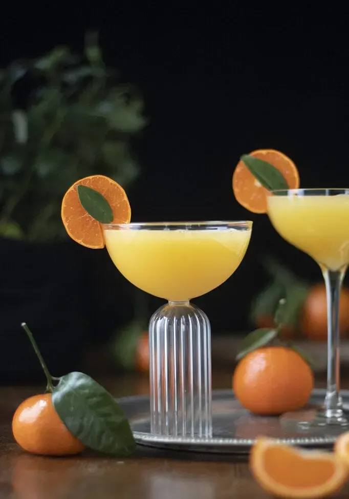 una elegante copa cupé llena de un cóctel de naranja y adornada con una rodaja de mandarina.' loading='lazy' title=
