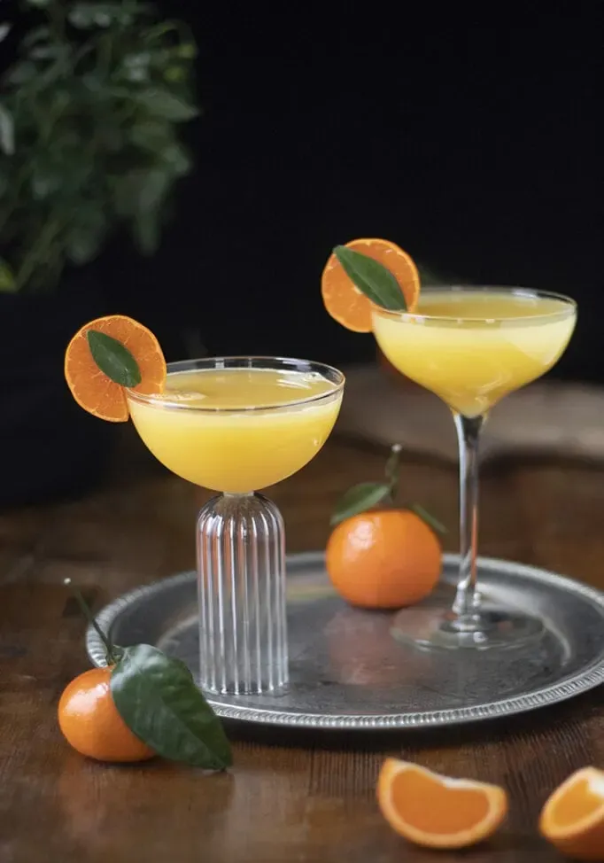 dos copas de cóctel llenas de una bebida de color naranja pálido en una bandeja de plata.' title=