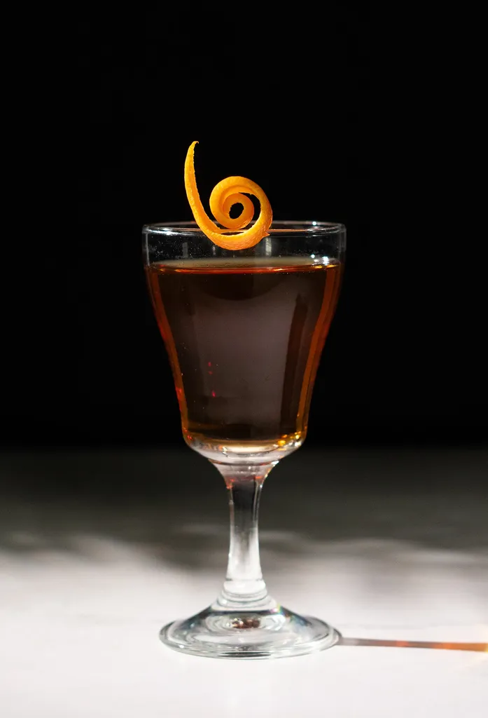 붉은 갈색 액체로 채워지고 작은 오렌지 껍질로 장식된 작은 칵테일 잔.' title=