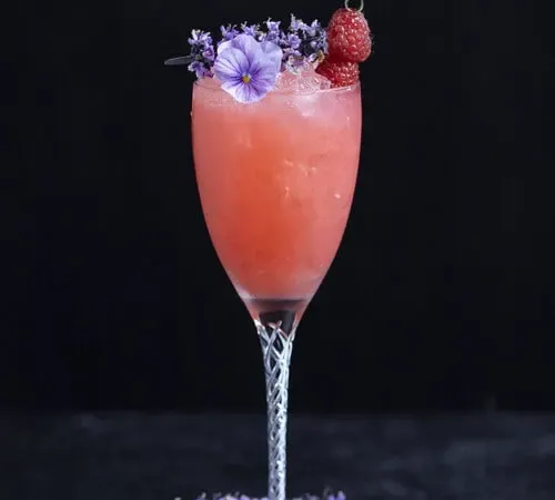 ein langstieliges Weinglas, gefüllt mit einem rosa Cocktail, garniert mit lila Blumen' title=