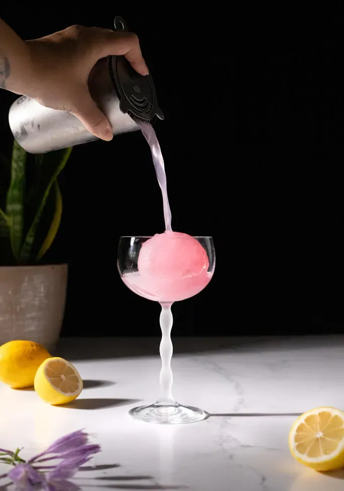 מזיגת משקה ורוד לכוס קופה מלאה בכדור קרח ורוד.' loading='lazy' title=