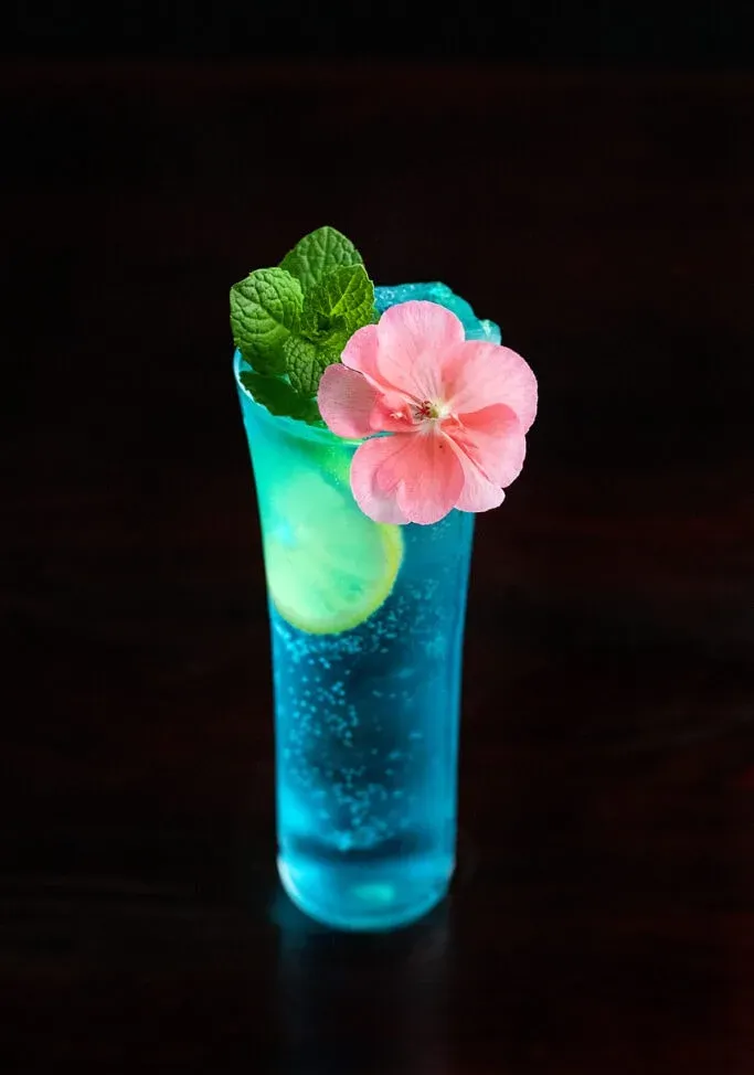 青いカクテルの上にピンクの食用の花。' title=