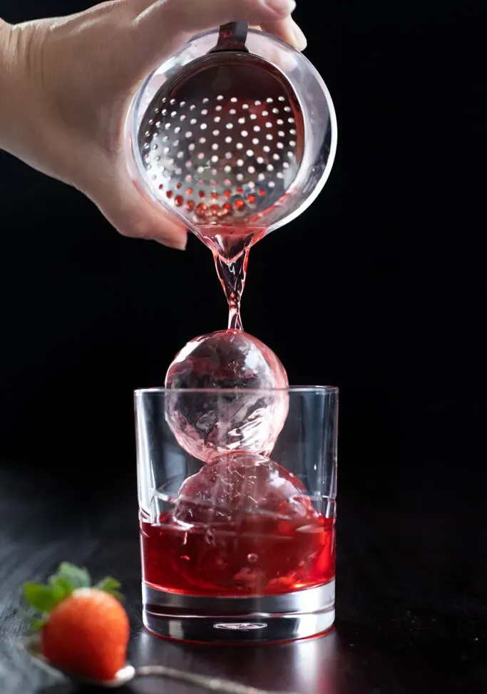 derramando uma bebida vermelha sobre duas grandes esferas de gelo transparente.' loading='lazy' title=