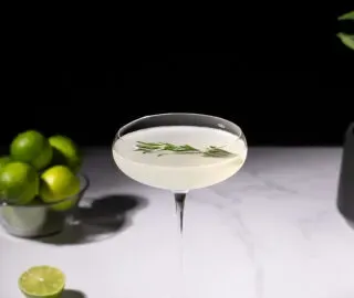 Ein Cocktail in einem hohen Stielglas mit einem Rosmarinzweig neben geschnittenen Limetten.' title=