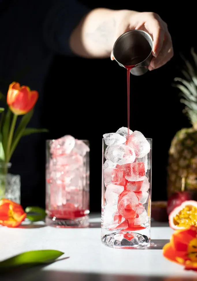 Gießen Sie roten Sirup aus einem Metallbecher in ein mit Eis gefülltes Longdrinkglas.' loading='lazy' title=