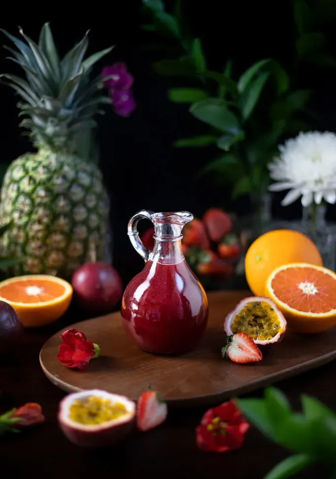 kleiner Krug mit dunkelrotem Sirup neben vielen tropischen Früchten.' title=