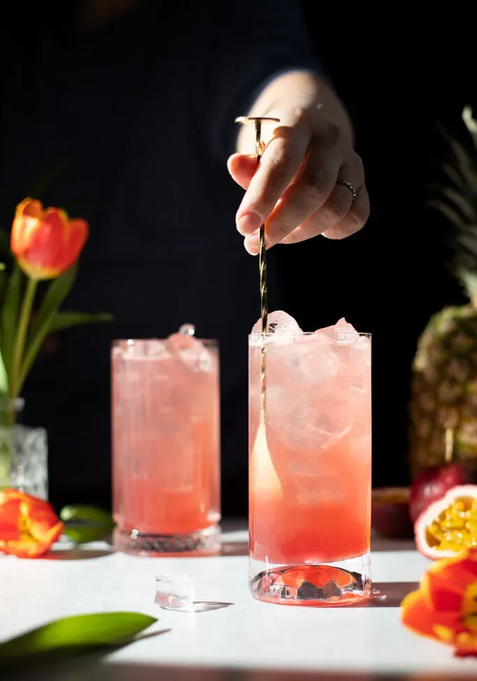 Hand rührt ein großes rosafarbenes Getränk mit einem goldenen Barlöffel um.' title=