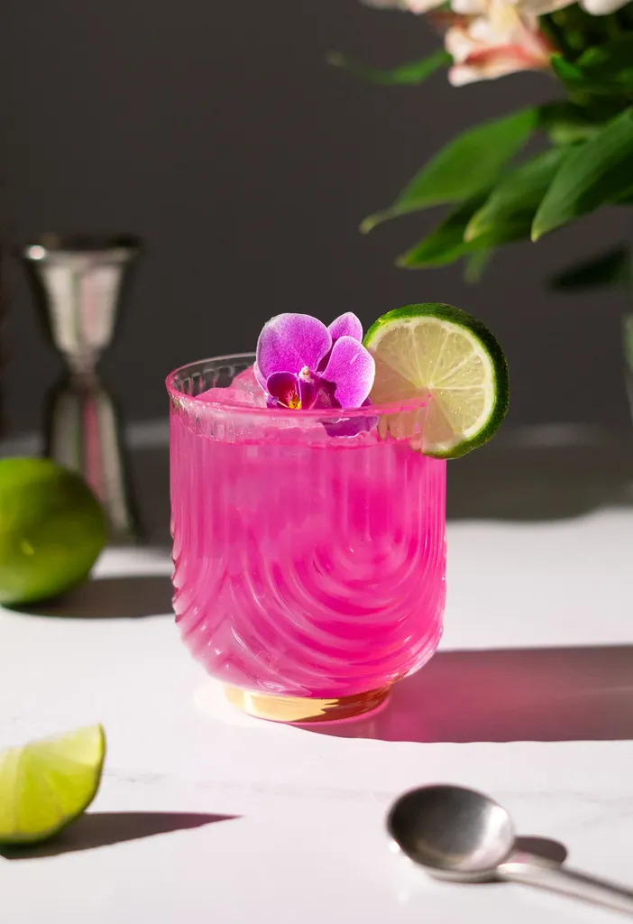 白い花とメタルジガーの隣の白いテーブルの上のロックグラスに入ったホットピンクの飲み物。' title=