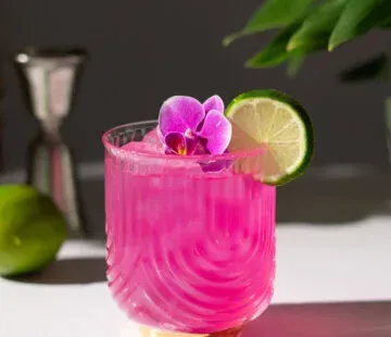 白い花とメタルジガーの隣の白いテーブルの上のロックグラスに入ったホットピンクの飲み物。' title=