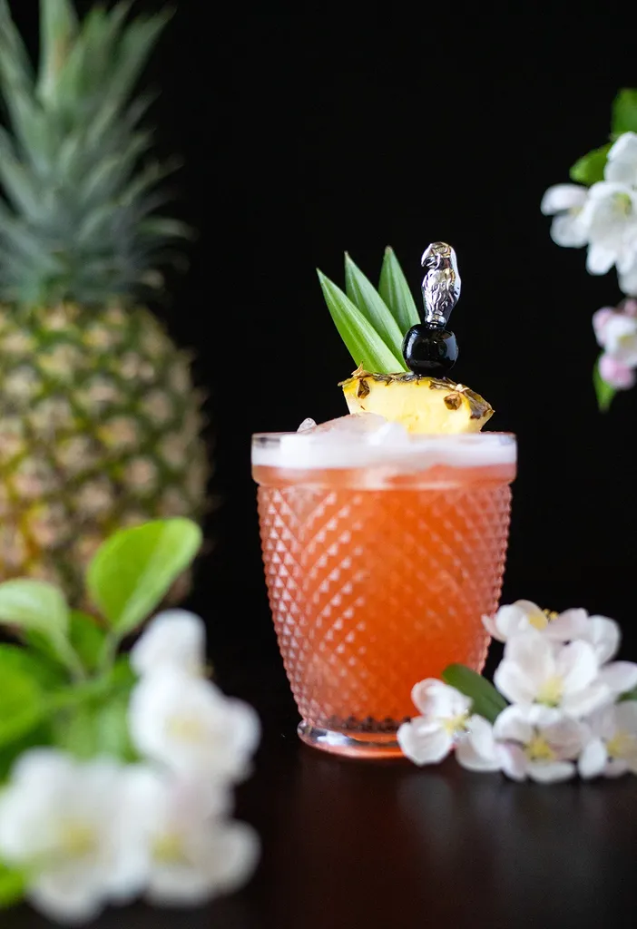 Rot-orangefarbenes Getränk in einem Steinglas neben einer Ananas und weißen Blumen.' loading='lazy' title=