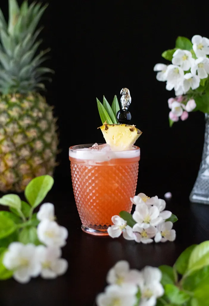 Rot-Orange-Getränk mit Ananasspalte neben weißen Blüten.' title=