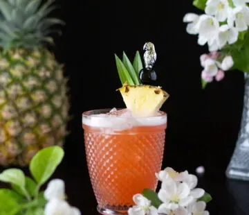 Rot-Orange-Getränk mit Ananasspalte neben weißen Blüten.' title=