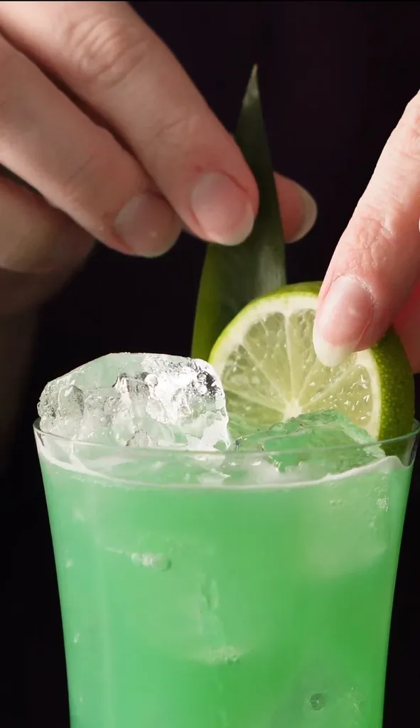 緑のカクテルにライムホイールとパイナップルの葉の飾りを置く手。' title=