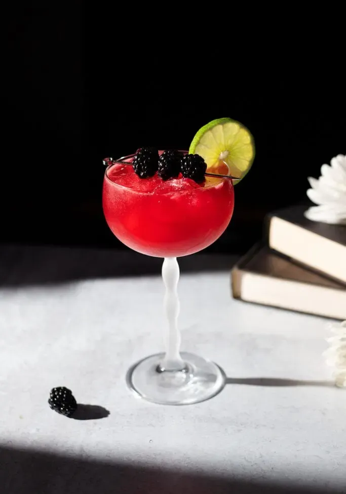 Leuchtend roter Cocktail in einem Stielglas, garniert mit Brombeeren und einem Limettenrad in der Sonne' title=