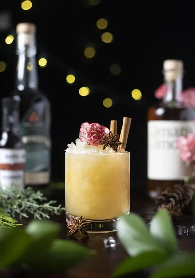 Cóctel de color amarillo pálido en un vaso bajo junto a botellas de alcohol.' title=