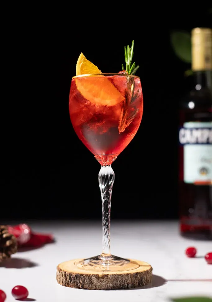 la bevanda rossa finita in un bicchiere di vino con fetta d'arancia e rosmarino.' title=