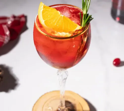 bicchiere di vino riempito con un cocktail rosso e guarnito con arancia e rosmarino.' title=