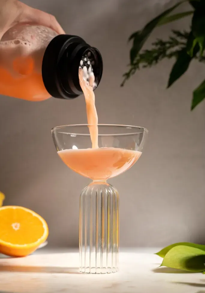 versando un liquido arancione dallo shaker in un bicchiere da coupé.' title=
