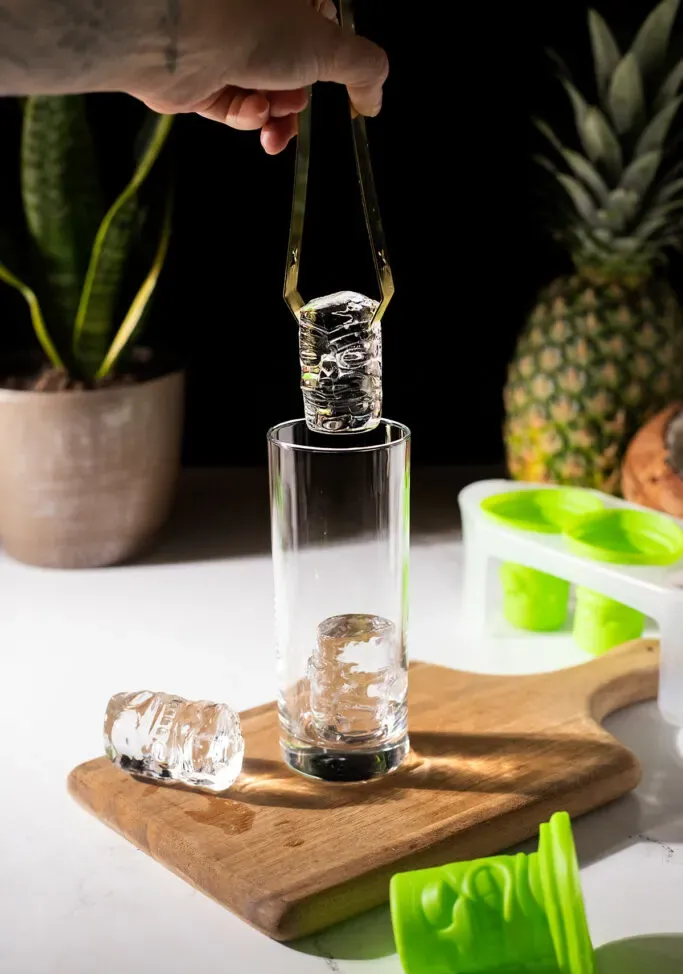 얼음 집게를 사용하여 투명한 티키 얼음 조각을 키가 큰 유리잔에 넣습니다.' title=