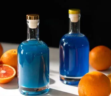 dwie szklane butelki wypełnione niebieskim płynem obok pokrojonych pomarańczy.' title=