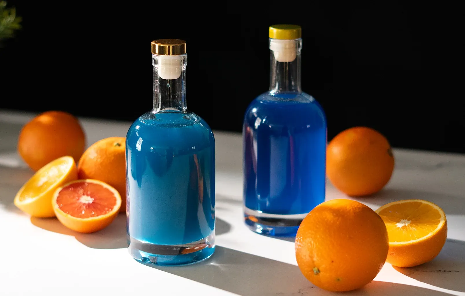 dwie szklane butelki wypełnione niebieskim płynem obok pokrojonych pomarańczy.' loading='lazy' title=