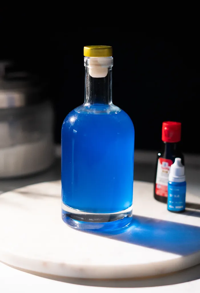 szklana butelka wypełniona jasnoniebieskim płynem obok niebieskiej butelki z barwnikiem spożywczym.' title=