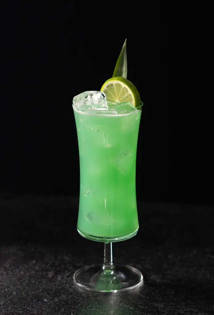 jasnozielony koktajl w szklance typu huragan ozdobiony kółkiem limonki i liściem ananasa.' title=