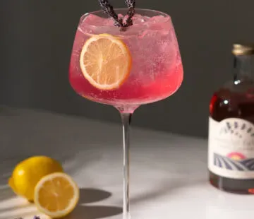Rosafarbener Cocktail neben Zitronen und einer Flasche Lavendelsirup.' title=