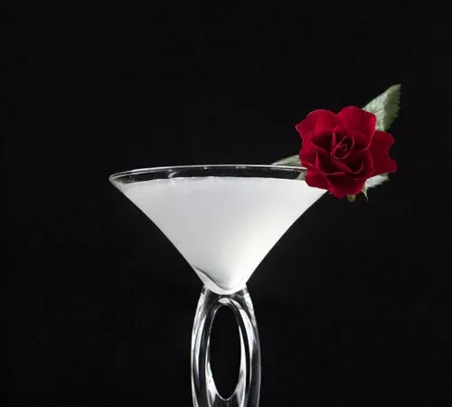 weiße Flüssigkeit in einem Martiniglas mit einer roten Rose am Rand.' title=