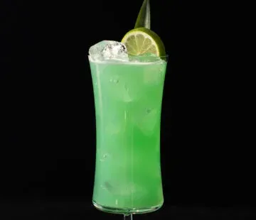 קוקטייל ירוק עז בכוס הוריקן מעוטר בגלגל ליים ועלה אננס.' title=
