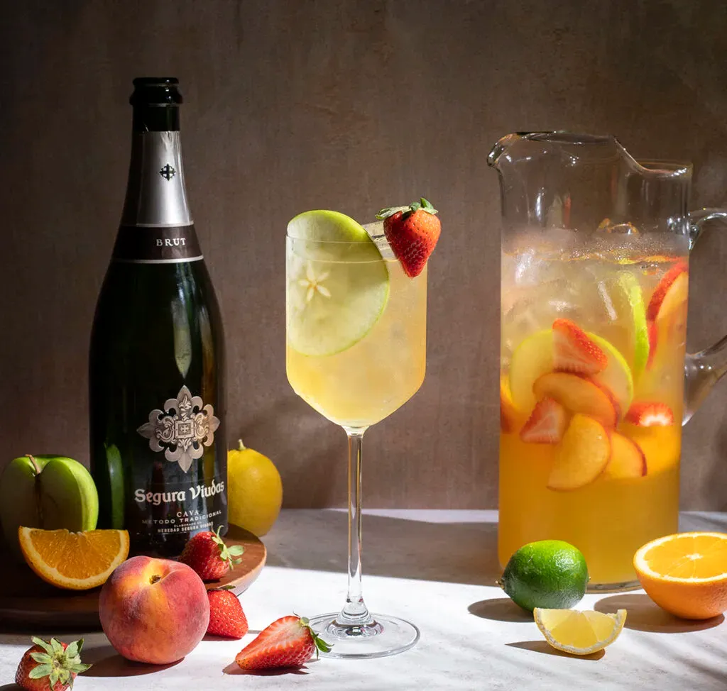 vysoký pohár na víno naplnený žltou sangriou obklopený ovocím a fľaša cava.' title=