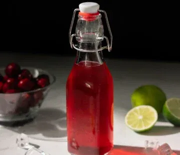 בקבוק נוזל אדום ליד קערת זכוכית קטנה של דובדבנים אדומים כהים.' title=