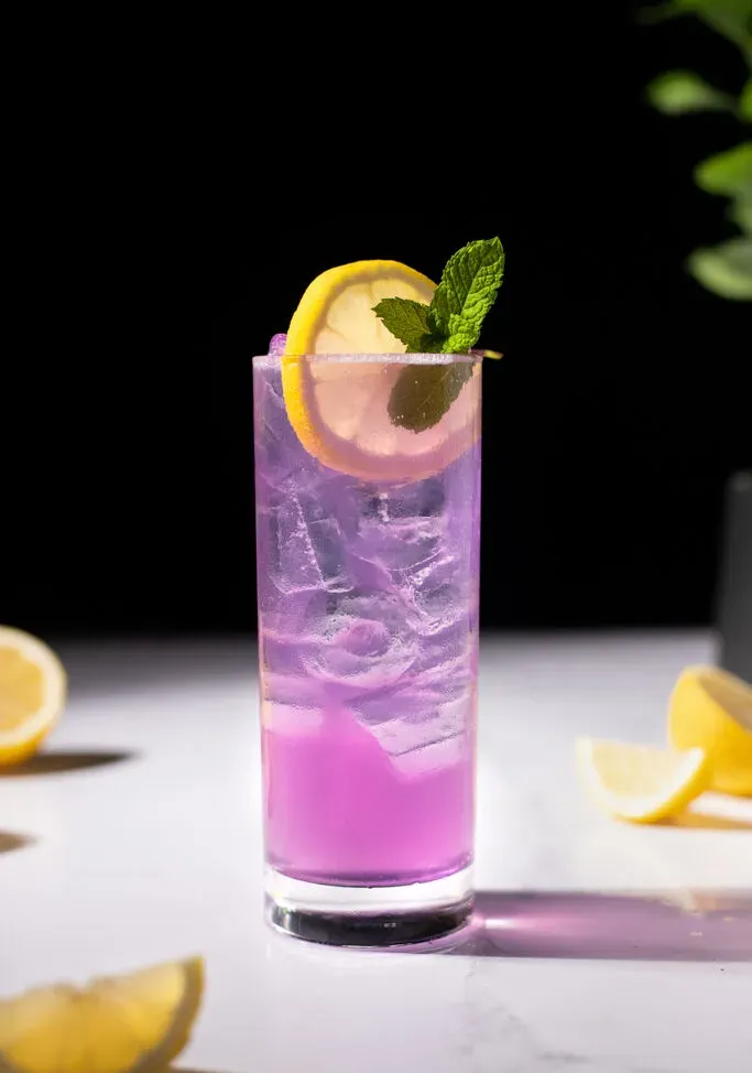 una bebida morada en un vaso Collins con una rodaja de limón y menta.' title=